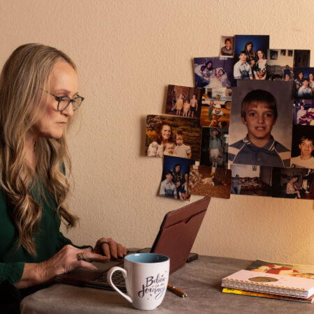 A woman with long blonde hair and glasses sits at a laptop. A collage of photographs is on the wall beside her. The photographs are of her son at various stages of his life.