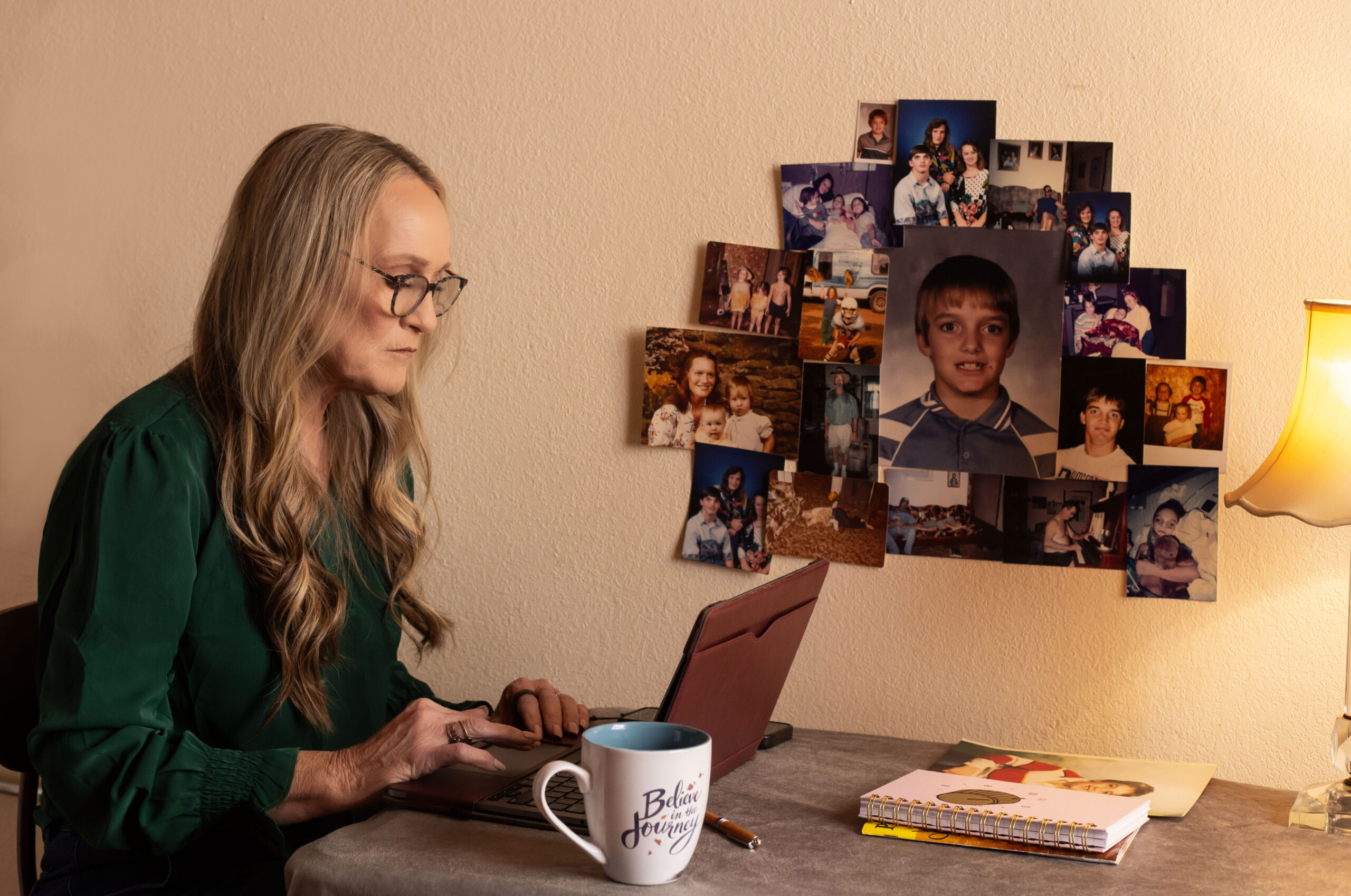 A woman with long blonde hair and glasses sits at a laptop. A collage of photographs is on the wall beside her. The photographs are of her son at various stages of his life.