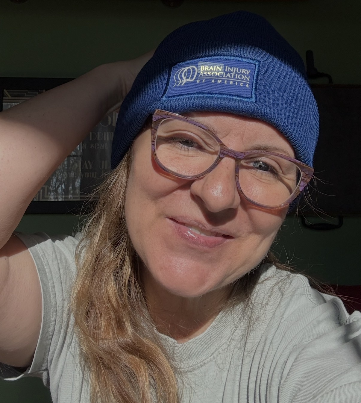 A woman with blonde hair and glasses is smiling. She is wearing a blue beanie with the Brain Injury Association of America logo on the front.