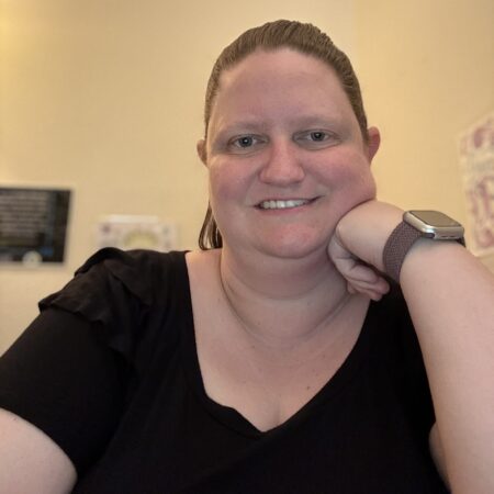 A smiling woman wearing a black t-shirt and a wristwatch, with reddish-brown hair in a ponytail.